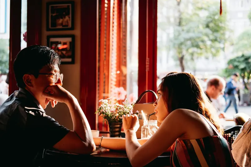 Couple sitting across from each other at a café table by a window, warm afternoon light