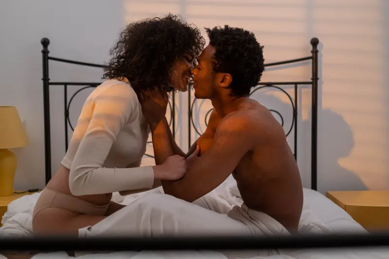 Couple in bed leaning close together, playful moment, soft morning light through blinds