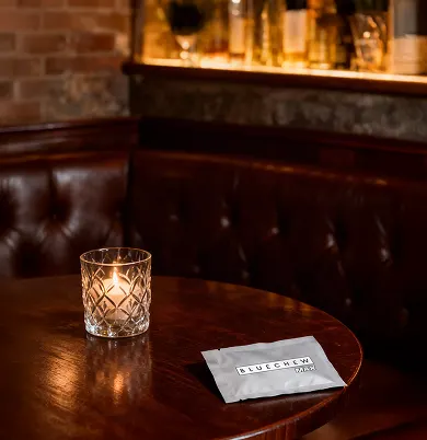 Gray BlueChew packet resting on a dark wooden table beside a lit candle in a cocktail lounge setting.