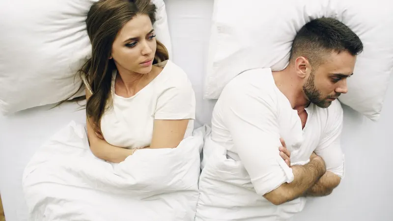 Couple lying back-to-back in bed, both looking distressed or disconnected