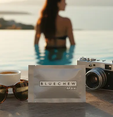 Woman standing in an infinity pool at sunset, with a gray BlueChew MAX packet, sunglasses, and a camera in the foreground.