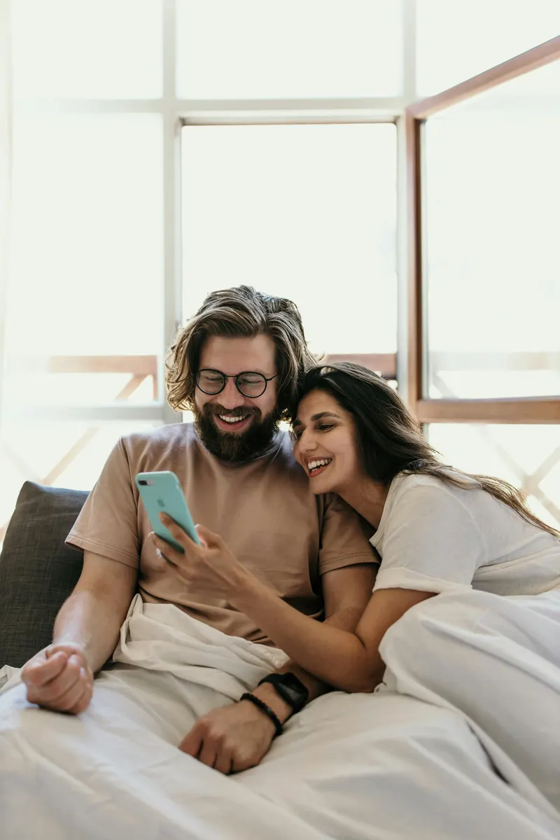Couple sitting in bed together smiling while looking at a phone.