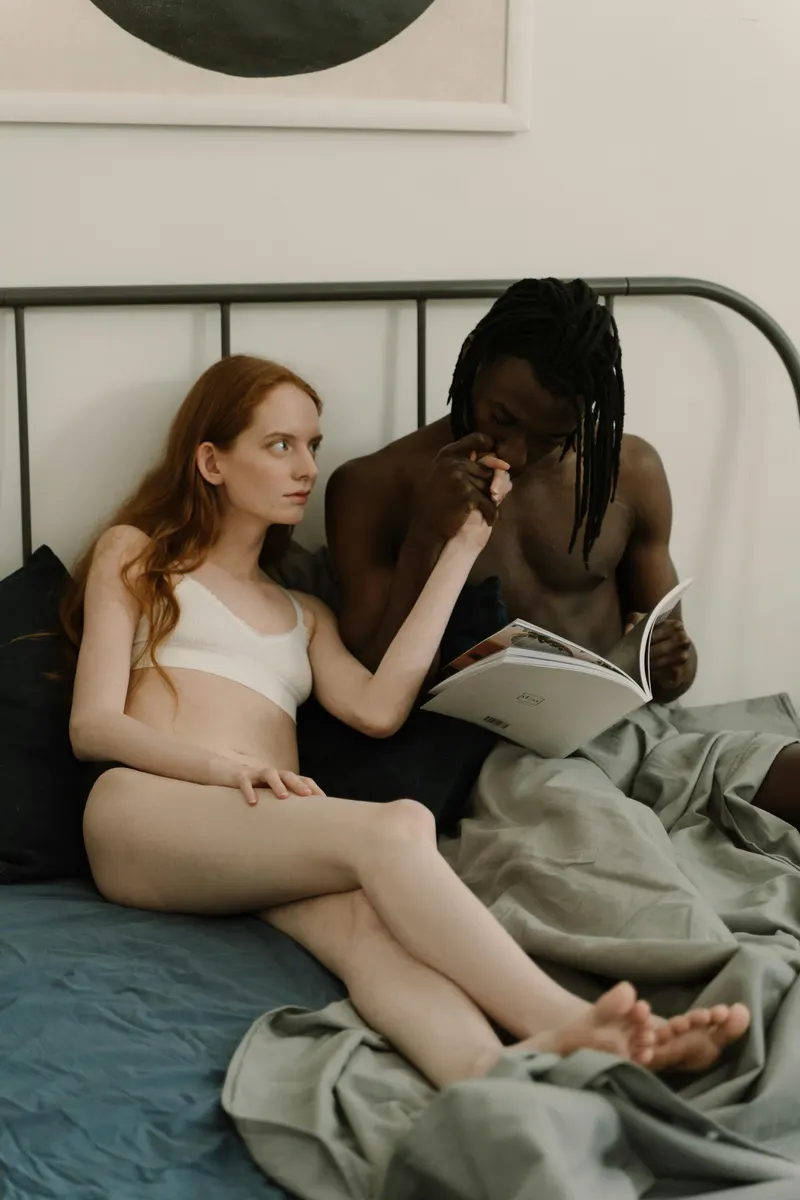 Interracial couple relaxing together in bed after intimacy, woman resting against her partner as he reads.