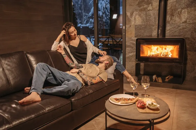 Couple relaxing on a leather couch by a fireplace with wine and food on the table, cozy winter setting