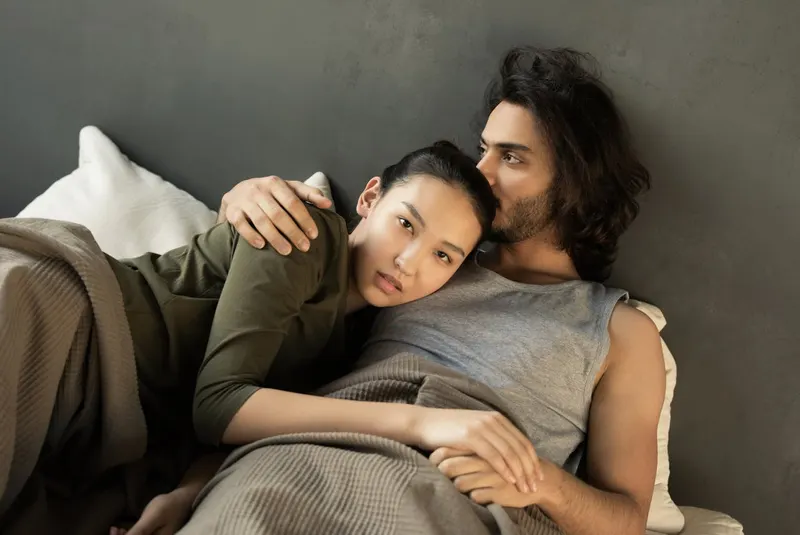 Couple embracing closely in bed, woman resting against man's shoulder, muted earth-toned bedding