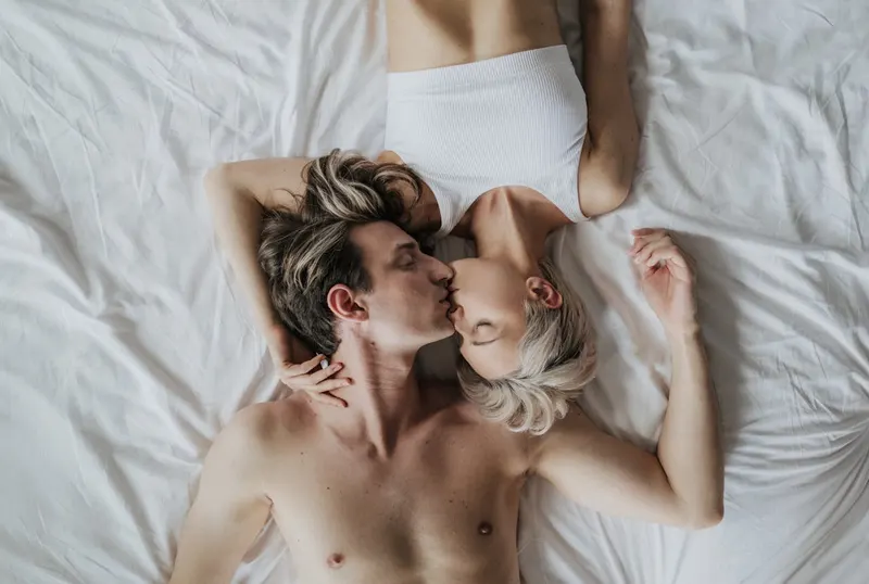 Couple lying on white bed sheets kissing, overhead view, playful morning moment