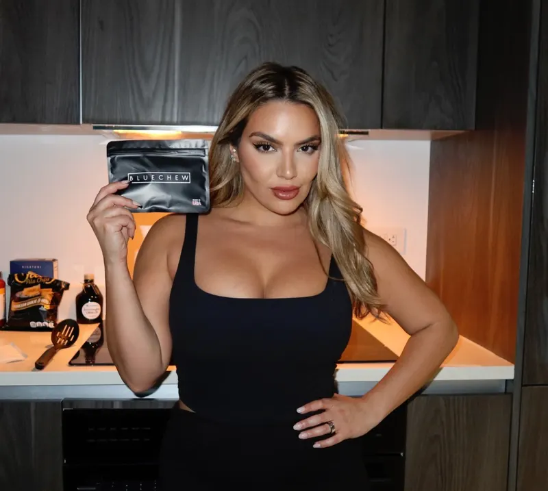 Woman in a black tank top standing in a modern kitchen, holding up a gold BlueChew packet.