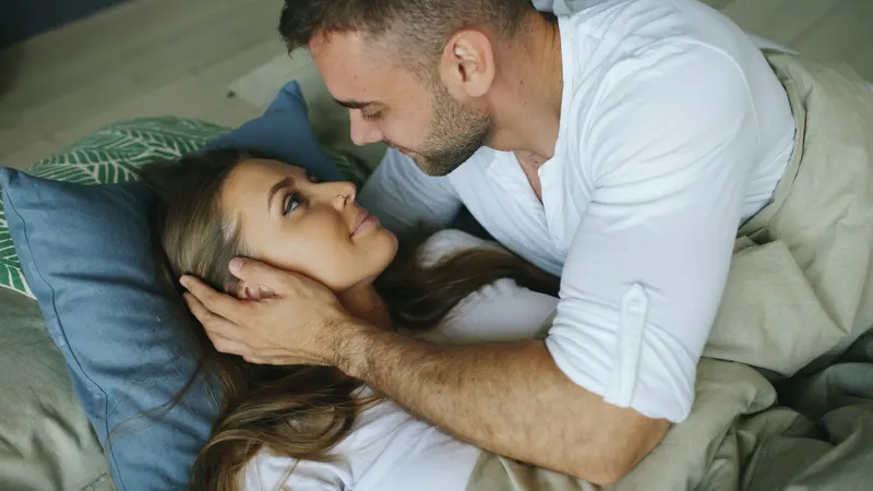 Couple embracing intimately kneeling on a bed near a bright window, moody vintage-style room