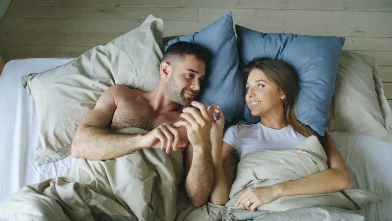 Couple lying in bed smiling at each other, holding hands under the covers.