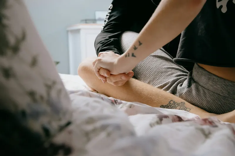 Close-up of a couple lying in bed holding hands, tattoos visible on their arms.