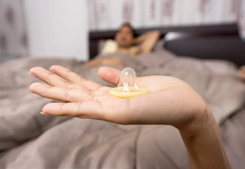 Open palm holding a condom in bed, partner relaxing in soft-focus background