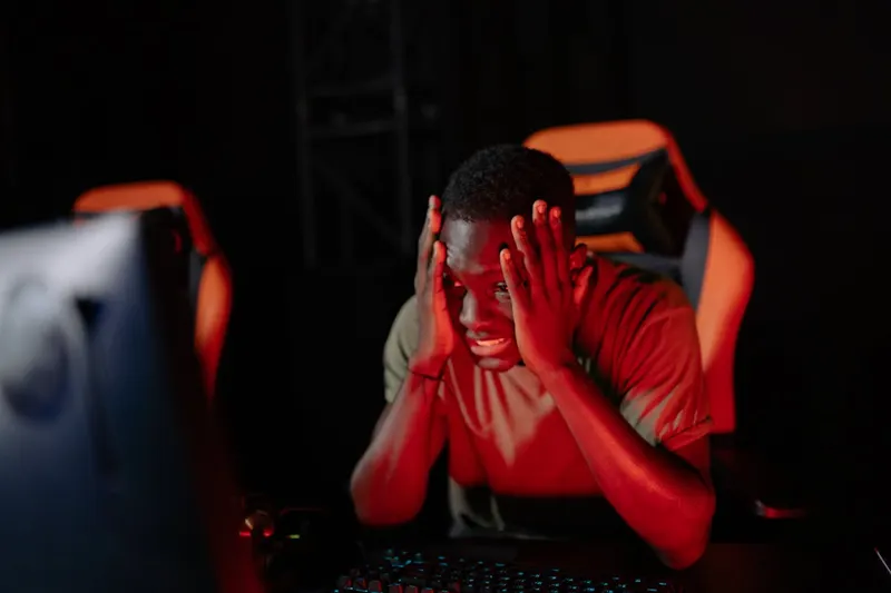 Man sitting at a gaming desk holding his head in his hands, dramatic red lighting, multiple monitors in background