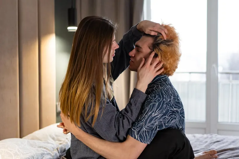 Couple embracing playfully in bed, morning light through window, woman ruffling man's hair