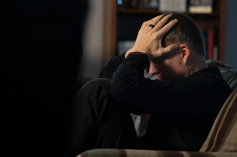 Man sitting on a couch holding his head in distress, dim lighting, bookshelf in background
