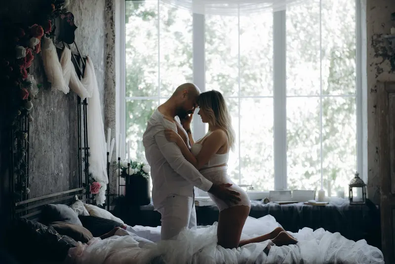 Couple embracing intimately kneeling on a bed near a bright window, moody vintage-style room