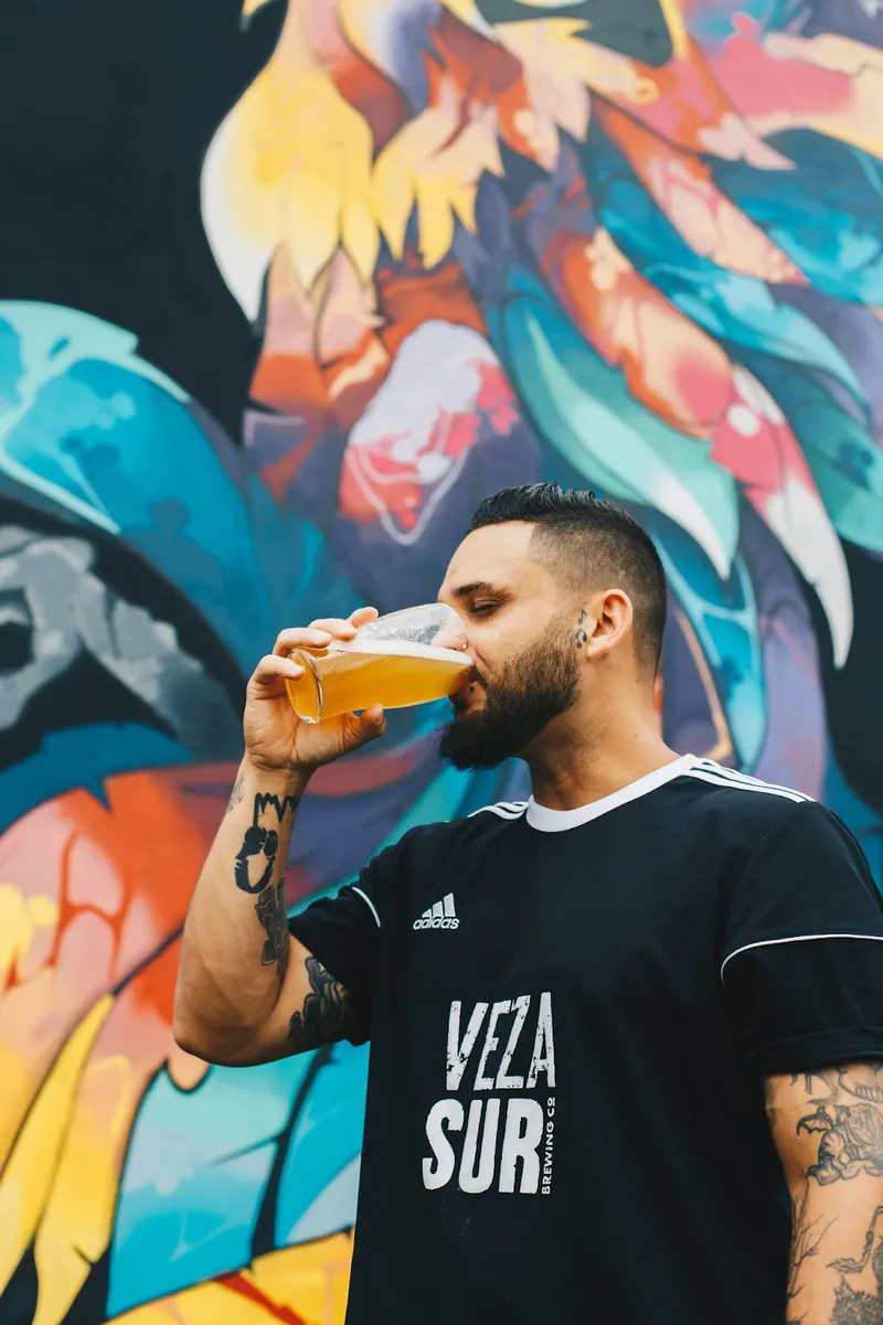 tattooed young man drinking a beer in front of a mural