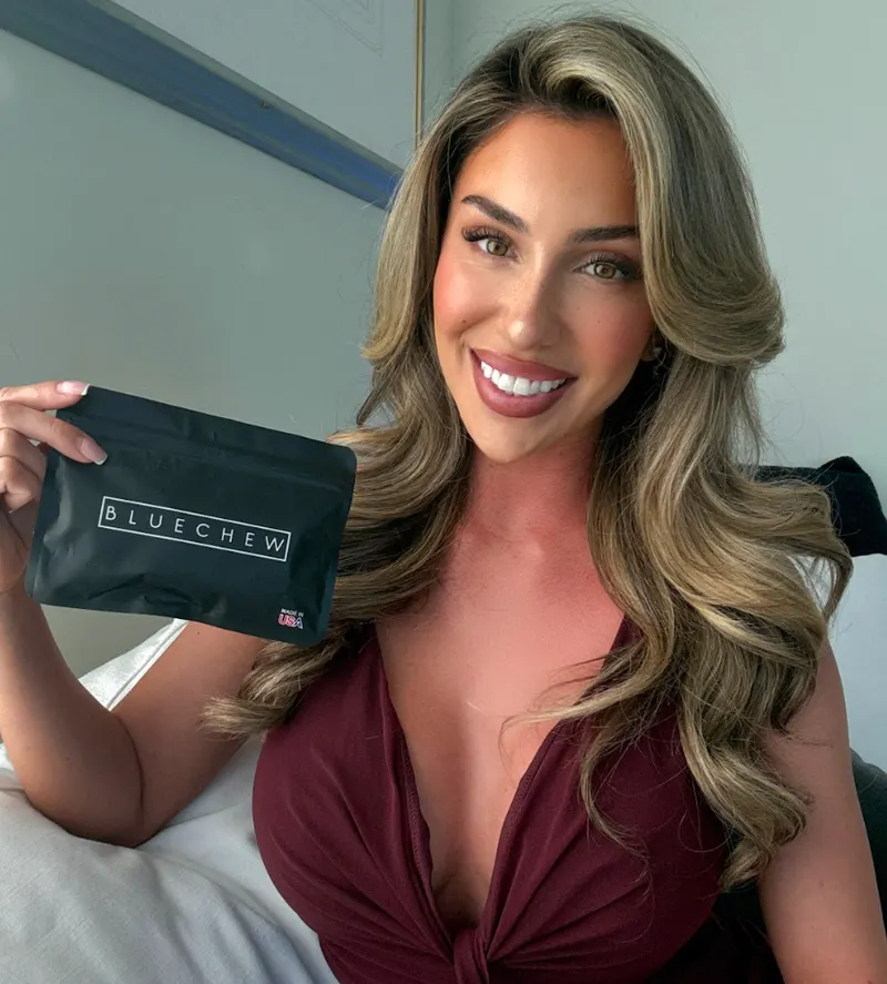 Smiling woman in a burgundy top holding a black BlueChew case, sitting on a bed