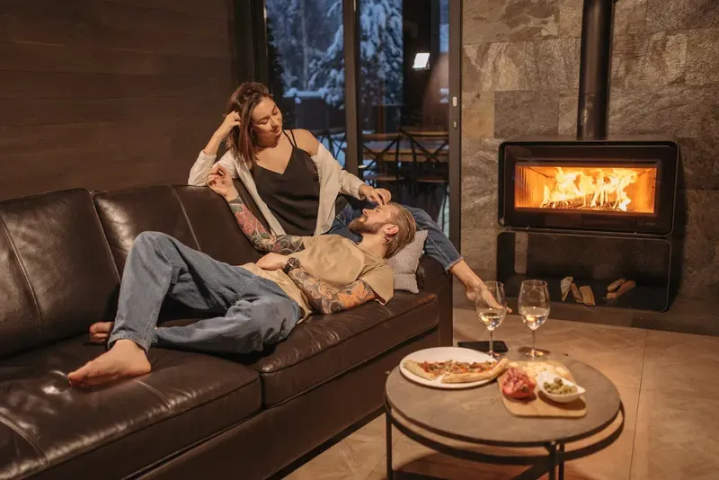 Couple relaxing on a leather couch by a fireplace with wine and food on the table, cozy winter setting