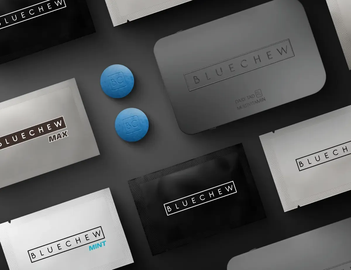 Diagonal close-up of black, white, and gray BlueChew MAX/MINT packets with two blue tablets on top.